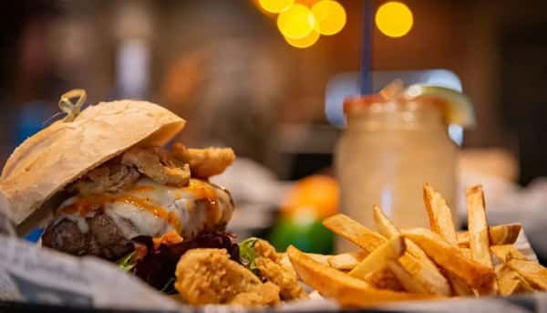 Cheeseburger topped with crispy onion rings and hot sauce, served with fries and a cocktail in a mason jar.