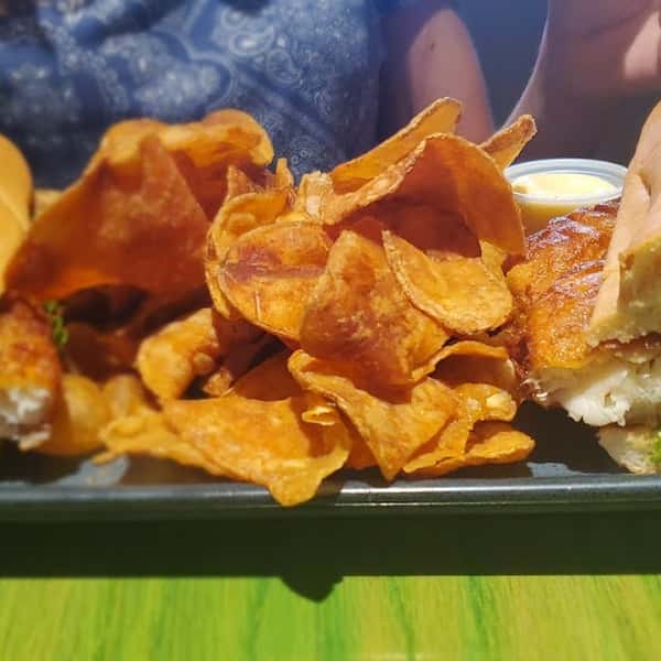 Crispy fish sandwich with a side of house-made potato chips and dipping sauce on a black plate.