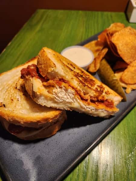 Grilled chicken sandwich with crispy chips, pickle spear, and a side of ranch dressing on a black rectangular plate.