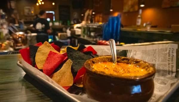 Tri-color tortilla chips served with a bowl of melted queso dip in a cozy bar setting.