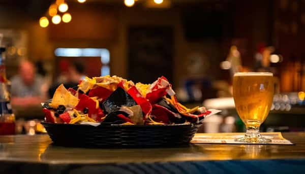 Basket of tri-color nachos topped with melted cheese, olives, and chicken, served with a pint of beer at the bar.