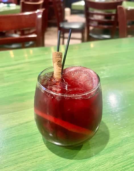 Glass of red sangria with ice, garnished with a lemon slice and cinnamon stick, served with black cocktail straws.