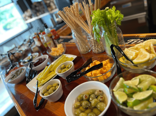 J.D.'s Bar-B-Que Bloody Mary garnish bar featuring green olives, cheddar cheese cubes, celery stalks, pickles, and fresh lemon/lime wedges on a wooden counter.