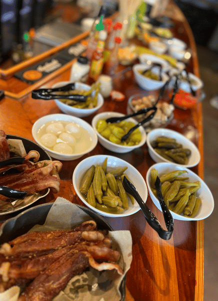 J.D.'s Bar-B-Que counter featuring platters of bacon, pickled okra, pearl onions, and peppers served buffet-style.