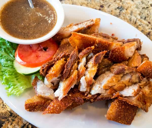 A serving of crispy fried pork belly slices (Lechon Kawali) with golden crackling skin, served with fresh tomato, greens, and a savory dipping sauce.
