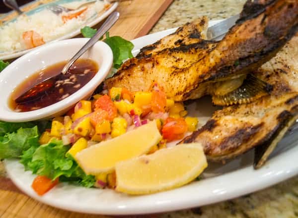 a whole grilled fish with crispy golden skin, served with lemon wedges, fresh salsa, and dipping sauce, creating a vibrant and flavorful seafood dish.