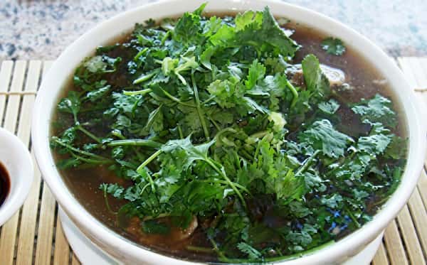A steaming bowl of pho soup with rich broth, tender meat, and fresh herbs generously topped with cilantro and greens.