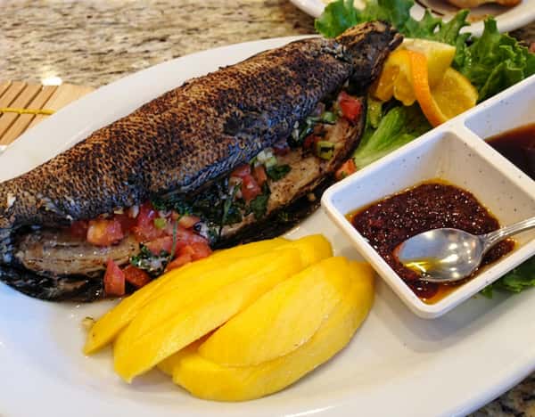 grilled fish with crispy, charred skin, served on a platter with fresh mango slices and a small bowl of dark dipping sauce. The dish is garnished with colorful vegetables and herbs, giving it a vibrant tropical presentation.