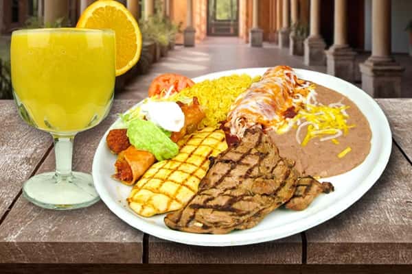 A hearty Mexican-style plate featuring sliced grilled steak, seasoned rice topped with shredded carrots, charred corn on the cob, and a side of creamy guacamole. The dish is vibrant and generous, served on a rustic wooden table with a drink in the background, evoking a warm, casual dining experience.