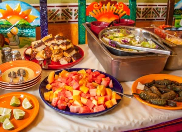 A colorful buffet spread featuring fresh-cut fruit in the foreground, alongside a variety of Mexican dishes including grilled items and a large tray of prepared food. The setup is vibrant and abundant, with warm tones, traditional decor, and a welcoming, festive atmosphere perfect for group dining or catering.