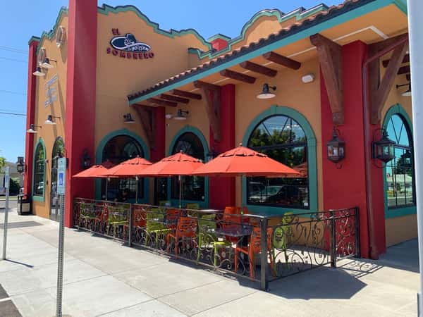 A vibrant Mexican restaurant exterior featuring warm red stucco walls, decorative trim, and a tiled roof. The outdoor patio is lined with colorful railings and shaded by bright umbrellas, creating a lively and inviting space for open-air dining.