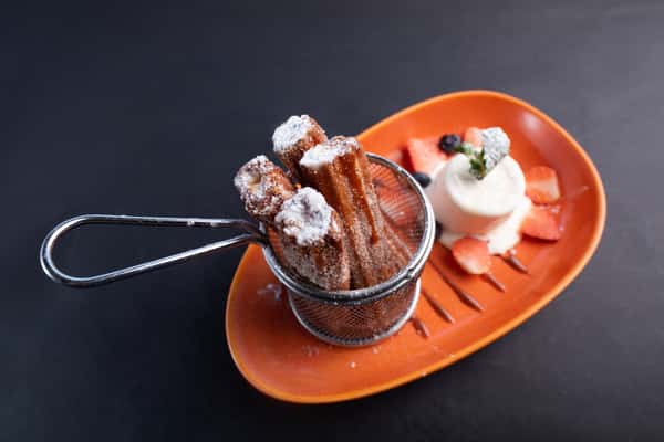 crispy churros dusted with sugar served with chocolate dipping sauce and whipped cream on an orange plate