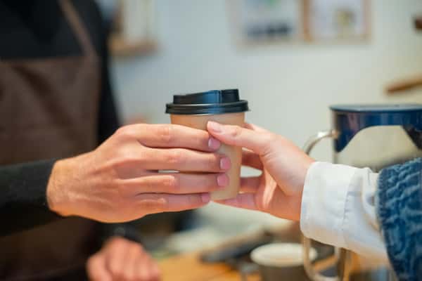 Barista handing a brown takeaway coffee cup with black lid to a customer inside a cozy café.