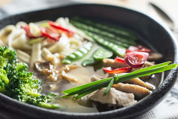 Bowl of noodle soup with sliced chicken, broccoli, snap peas, baby corn, cherry tomatoes, red chili, and green onions in broth.