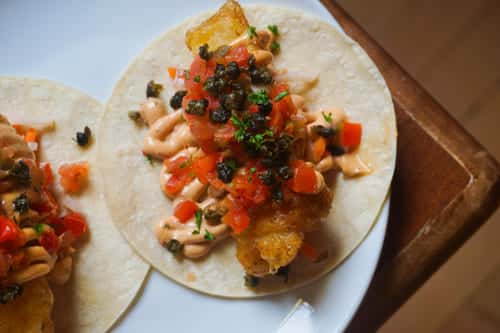 Fish taco topped with crispy fried fish, diced tomatoes, chipotle aioli, and capers on a soft flour tortilla—perfect for Baja-style cuisine.