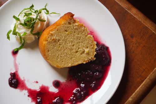 Moist slice of vanilla bundt cake served with whipped cream, mint garnish, and a drizzle of blueberry compote on a white plate.