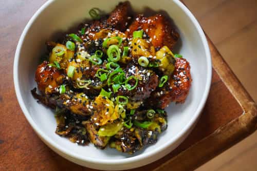Crispy glazed cauliflower and roasted Brussels sprouts garnished with sesame seeds and scallions in a white ceramic bowl, showcasing a savory vegan entrée.