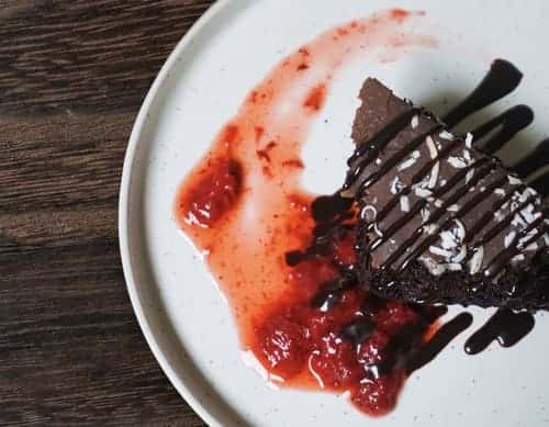 Decadent slice of chocolate cake topped with coconut flakes, drizzled with chocolate syrup and served with a strawberry compote.