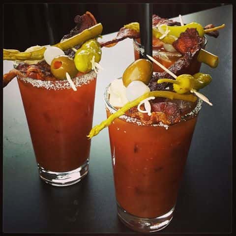 Three tall glasses with Bloody Mary cocktails topped with crispy bacon, pepperoncini, green olives and cheese bites