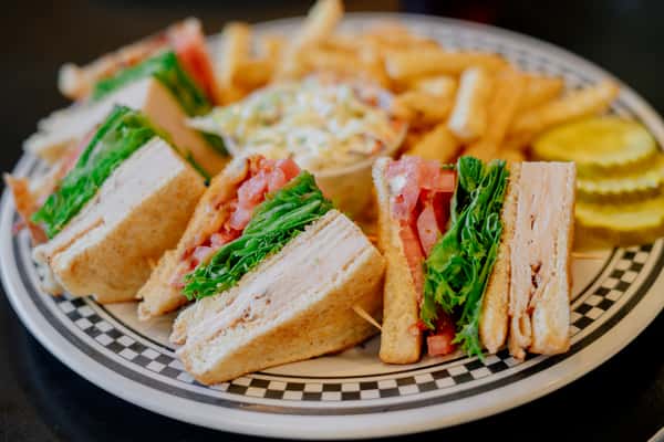 A freshly prepared club sandwich stacked with sliced turkey, crisp lettuce, tomato, and toasted bread, cut into quarters for easy serving. The sandwich is paired with golden French fries and crunchy pickles, creating a classic café lunch plate that's both hearty and satisfying.