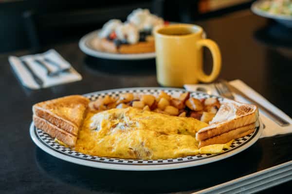 A classic café breakfast featuring a fluffy omelet topped with melted cheese, served with golden home fries and toasted bread. A warm cup of coffee sits nearby, creating a comforting, homestyle breakfast scene perfect for starting the day.