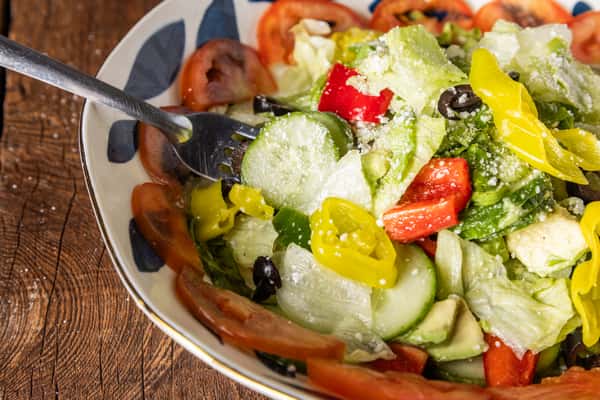 Fork mixing a fresh Italian garden salad with cucumbers, olives, banana peppers, lettuce, tomatoes, and grated Parmesan.