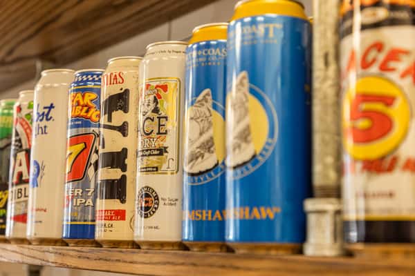 Craft beer and beverage cans displayed on a rustic wooden shelf, featuring assorted IPA, cider, and craft brew brands in a casual bar or restaurant setting. 🍺