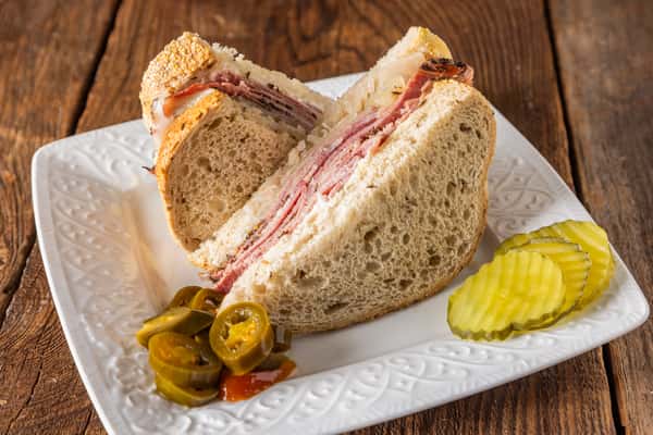 Classic deli sandwich with layered cured meats and melted cheese on artisan bread, served with sliced pickles and jalapeños.