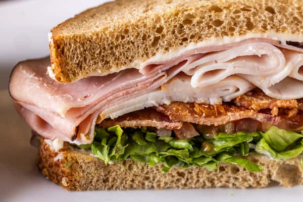 Turkey and ham club sandwich with crispy bacon, lettuce, tomato, and creamy sauce on toasted whole wheat bread, stacked deli-style for a hearty lunch sandwich.