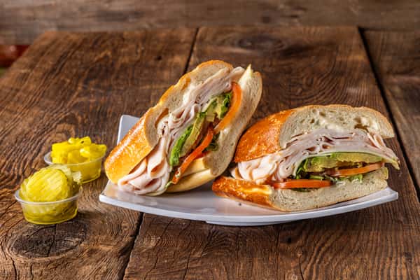 Turkey and avocado sandwich on artisan bread with tomato and lettuce served with pickles on a rustic wooden table.