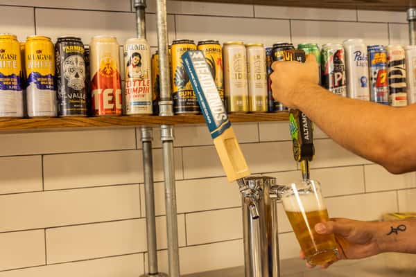 Bartender pouring draft craft beer from tap with a selection of canned craft beers displayed on a wooden shelf in a casual restaurant bar area.