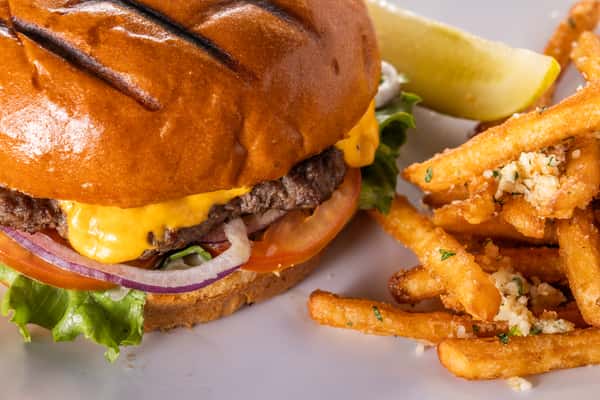 Grilled cheeseburger with melted cheddar, lettuce, tomato, and red onion on a toasted brioche bun served with crispy garlic-seasoned French fries and a pickle wedge on a white plate.