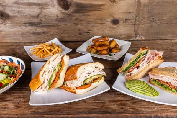 Assorted deli sandwiches, crispy fries, onion rings, and a fresh Italian salad arranged on plates over a rustic wooden table.