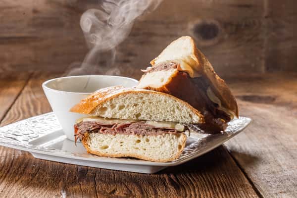 Hot roast beef sandwich on artisan bread served with steaming au jus dipping sauce on a rustic wooden table.