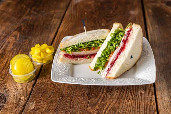 Turkey sandwich on white bread with cranberry spread, leafy greens, and sliced turkey served with pickles.