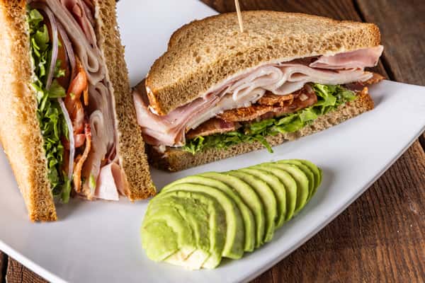 Turkey club sandwich on wheat bread with crispy bacon, lettuce, tomato, and sliced avocado served on a white plate.