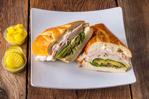Turkey and avocado sandwich on toasted artisan bread with fresh tomato, lettuce, and sliced turkey, served with pickles and pepperoncini on a white plate.