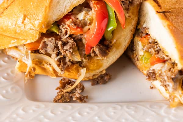 Philly cheesesteak sandwich with shaved steak, sautéed onions, melted cheese, and sliced red and green bell peppers on toasted bread, served on a white plate.