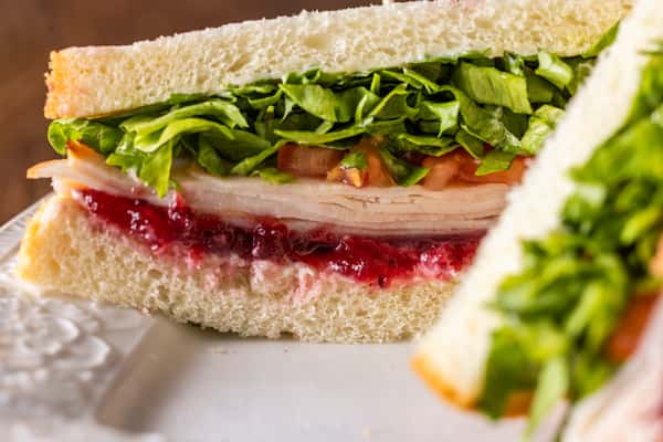 Turkey sandwich on soft white bread with cranberry spread, fresh leafy greens, and sliced tomatoes, served on a white plate in a classic deli-style presentation.