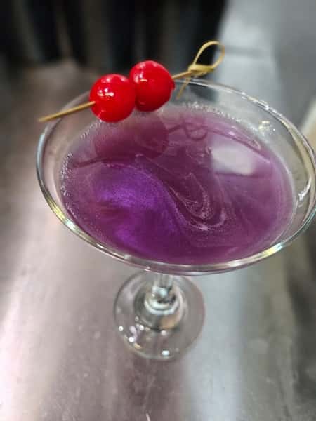 Vibrant purple cocktail in a martini glass garnished with two red cherries on a pick, sitting on a polished wooden bar.