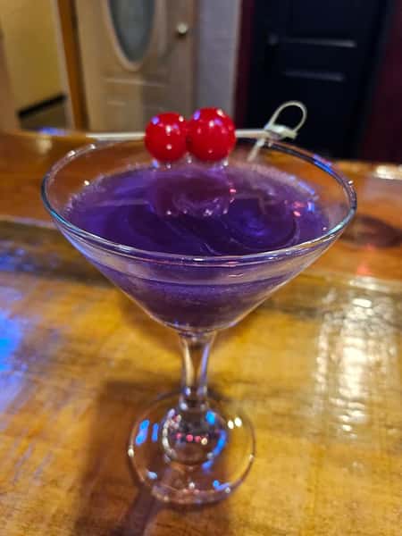 Vibrant purple cocktail in a martini glass garnished with two red cherries on a pick, sitting on a polished wooden bar.