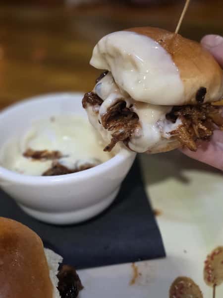 Pulled pork slider topped with melted white cheese, held above a bowl of dipping sauce on a plate.