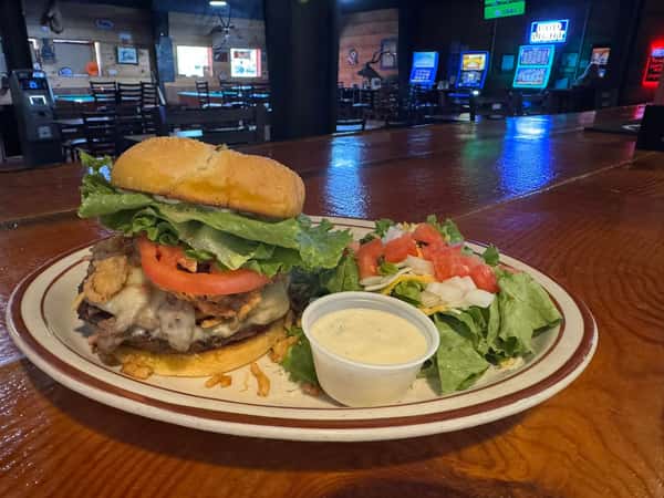 Cheeseburger with lettuce and tomato served with a side salad and ranch dressing on a plate at a bar counter.