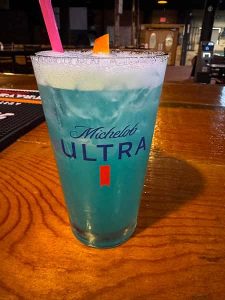 Tall glass of vibrant blue cocktail with a frothy top, garnished with an orange slice and a pink straw, served in a Michelob Ultra glass.