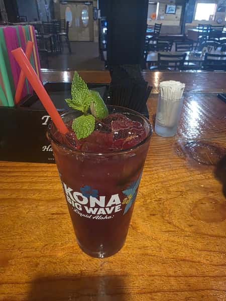 Purple iced cocktail in a Kona Big Wave glass, garnished with fresh mint and served with a red straw at the bar.