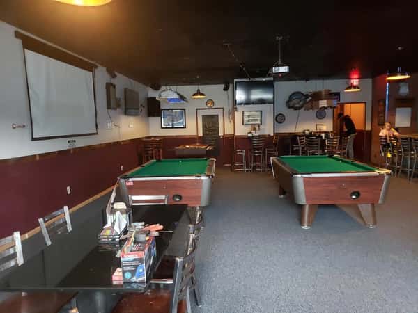 Bar interior with two pool tables, wall-mounted TVs, a projector screen, high-top seating, and board games on a nearby table.