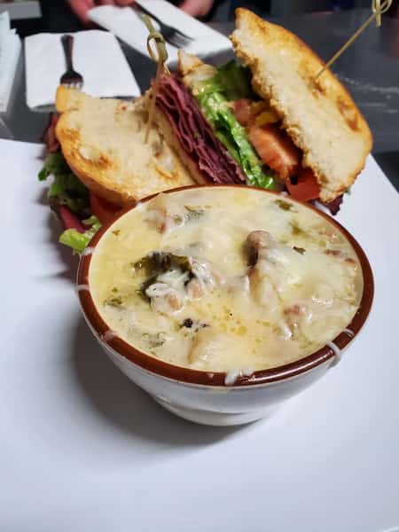 Hearty sandwich stacked with pastrami, lettuce, and tomato served with a creamy bowl of cheesy soup on a white plate.