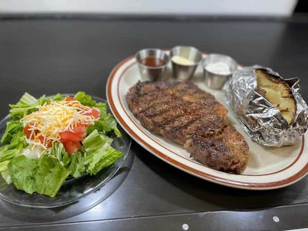 Grilled ribeye steak served with a baked potato, sauces, and a fresh side salad topped with shredded cheese and tomatoes.