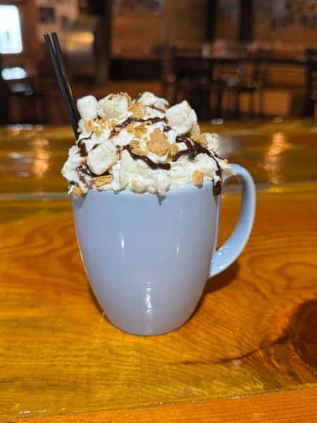 White mug filled with whipped cream, toasted marshmallows, crushed graham crackers, and chocolate drizzle, served on a wooden bar.