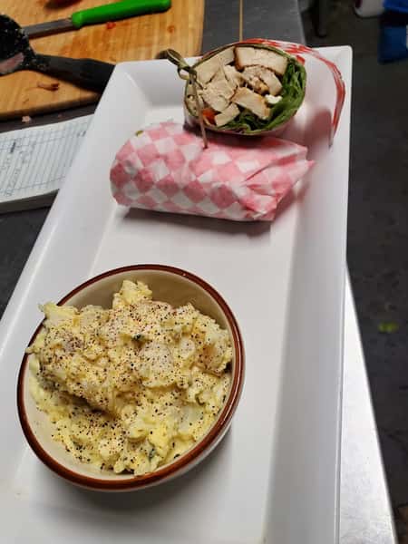 This meal features a grilled chicken wrap filled with fresh greens and vegetables, wrapped in pink-checkered paper, and served with a side of seasoned potato salad, garnished with black pepper.